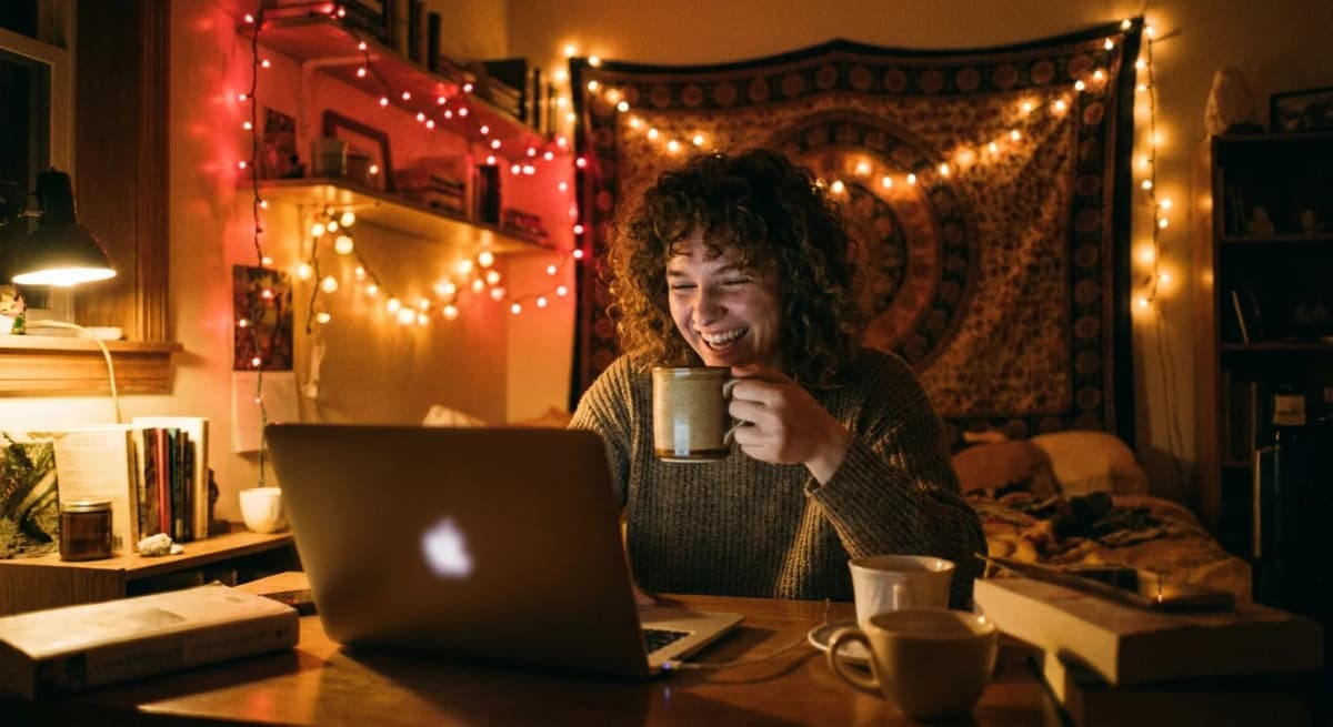 A young adult smiling and raising a drink toward a laptop webcam in a warm room strung with string lights, the screen turned away from the camera